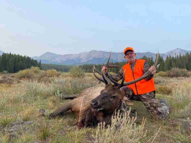 Muzzleloader hunter kneeling behind a wide-antlered bull elk in a sage meadow below the mountains of GMU 55, Colorado